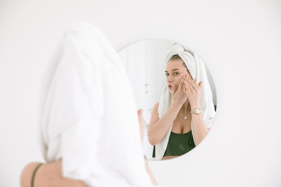 A woman applies face cream in the bathroom, reflecting in a mirror with a towel on her hea