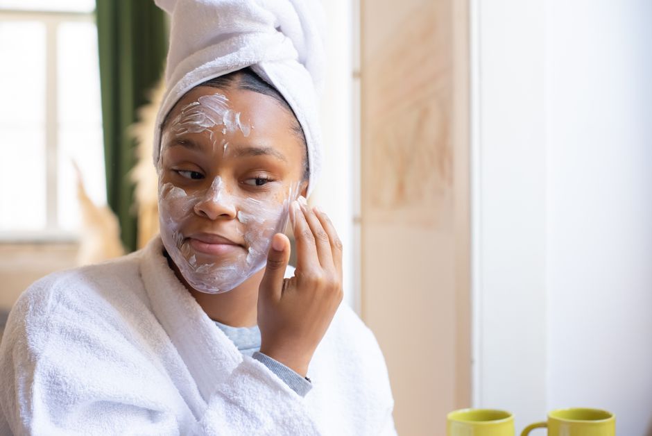 A young woman in a bathrobe applies face cream while relaxing indoors. Morning skincare ro