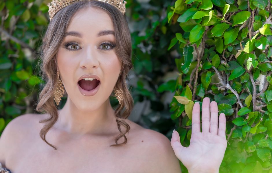 A young woman with a surprised expression and crown outdoors with vibrant greenery.
