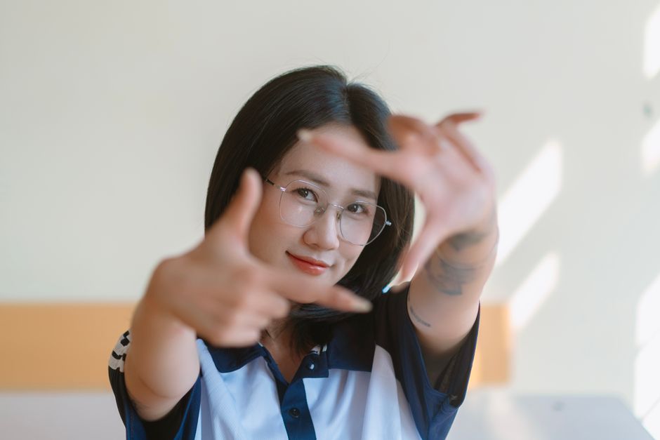 A young woman smiles as she frames her face with her hands in a sunlit room.