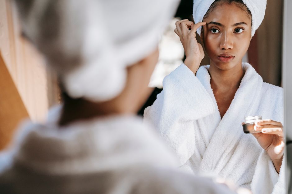 Young black female in white bathrobe and towel turban applying moisturizing cream on face while standing in bathroom and doing daily beauty procedure