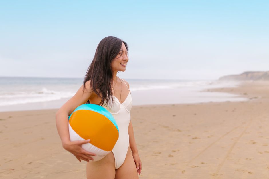 Asian woman in swimsuit holding beach ball on a sunny beach day.