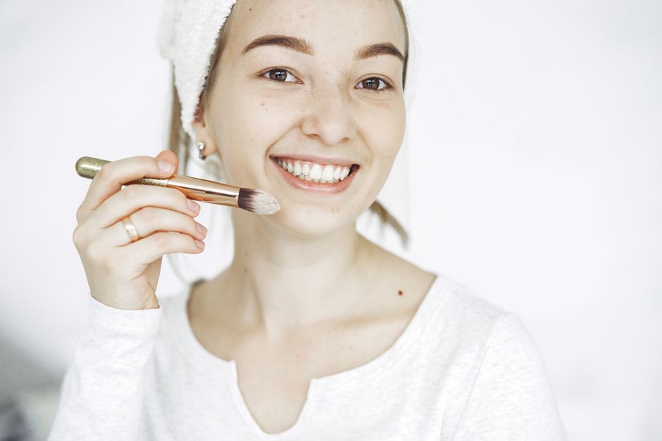 Young woman with a bright smile applying makeup indoors with a brush.