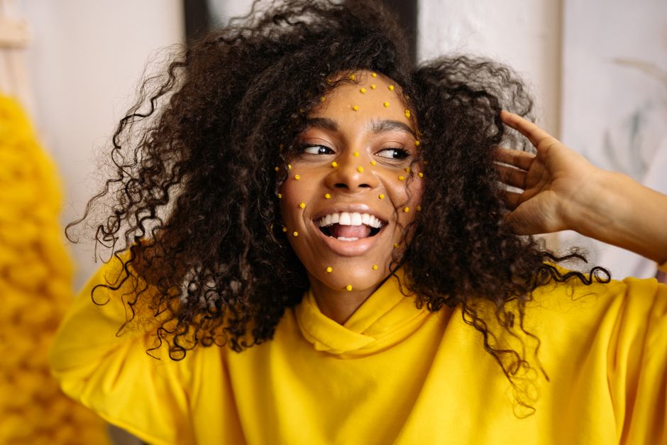 A happy woman with curly hair and art makeup in a vibrant yellow sweater, exuding joy and creativity.