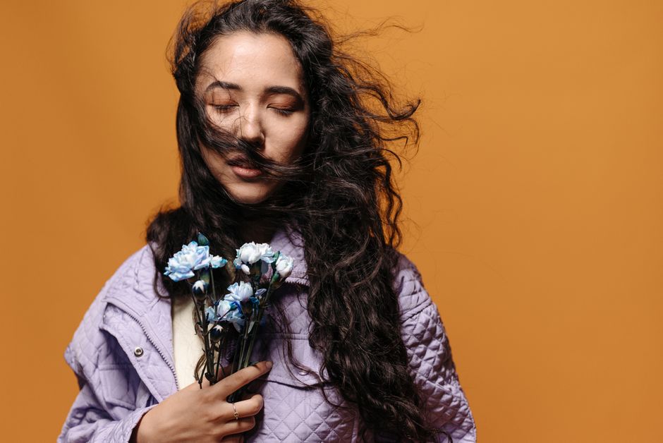 A woman with closed eyes holds flowers against an orange background, wearing a purple jacket.