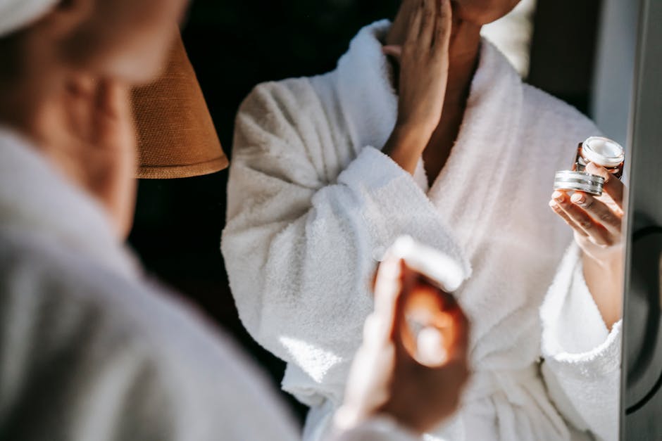 Crop anonymous female in white bathrobe applying moisturizing facial cream on face while standing in front of mirror in bathroom