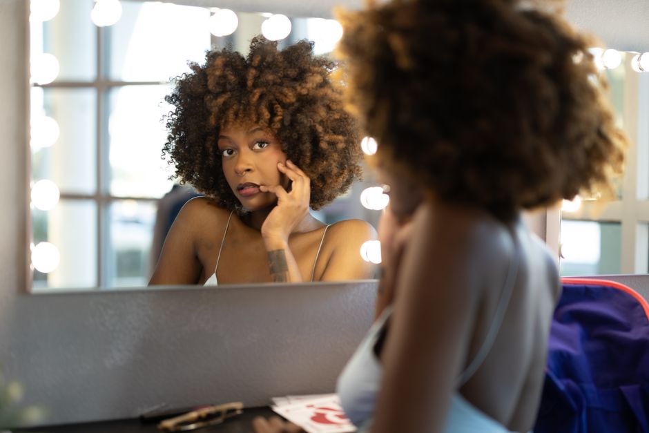 Woman with curly hair looking in a lit mirror, contemplating beauty and self-care.