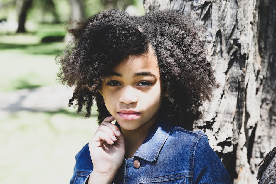 A thoughtful portrait of a young girl with curly afro hair leaning against a tree.