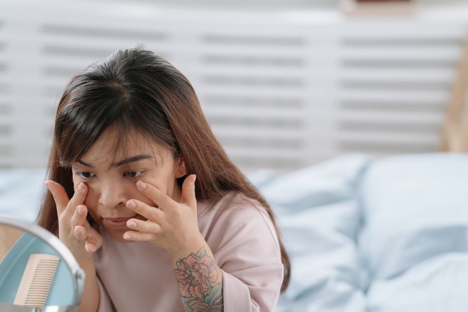 Asian woman applying skincare product in a bedroom setting with mirror reflection.