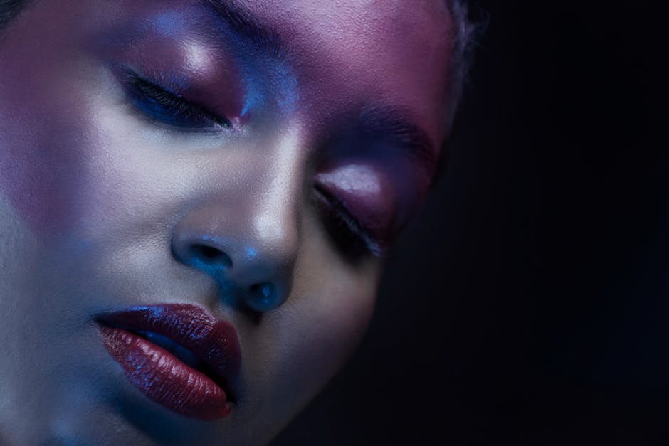 An artistic close-up of a woman's face with bold makeup and eyes closed, showcasing vibrant colors and a dark background.