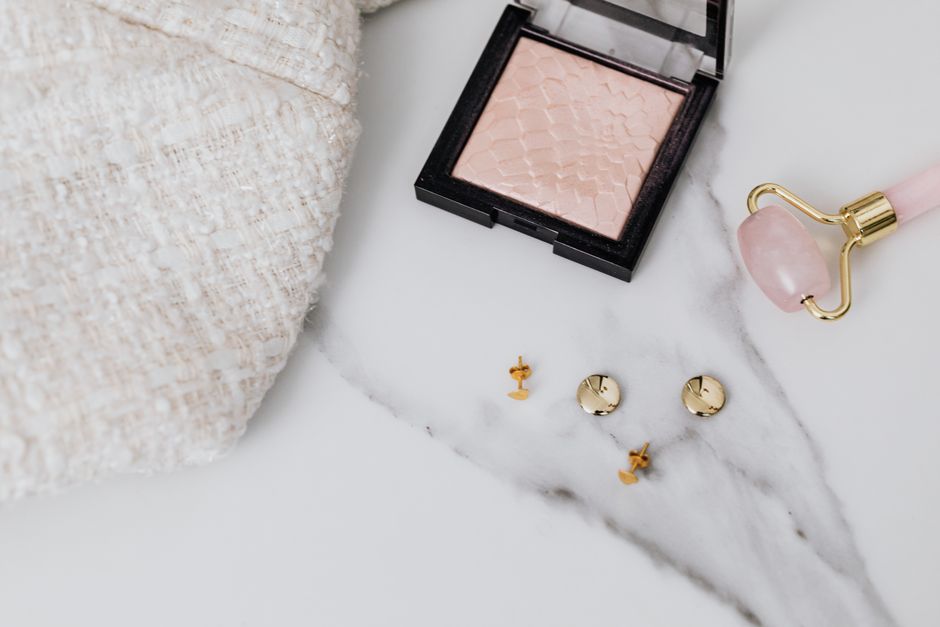 Flat lay of earrings, makeup powder, and jade roller on marble, displaying elegance.