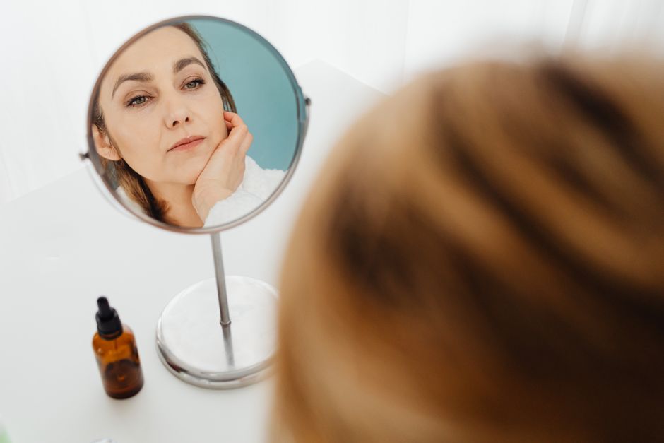 Adult woman examining her face in a mirror next to a skincare bottle.